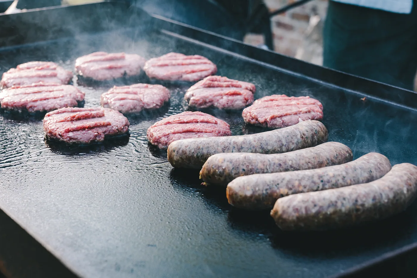 A&K beef patties and bratwurst cooking on a flat-top griddle