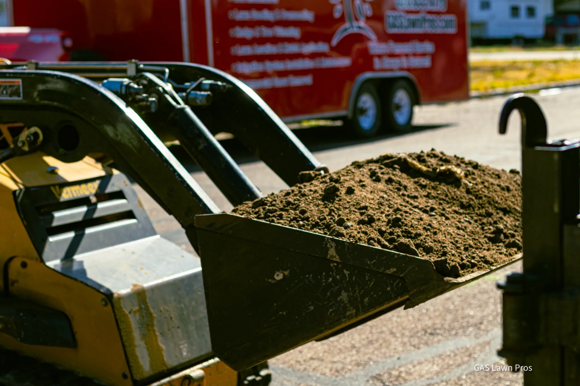 Loader bucket moving material beside a branded GAS Lawn Pros trailer