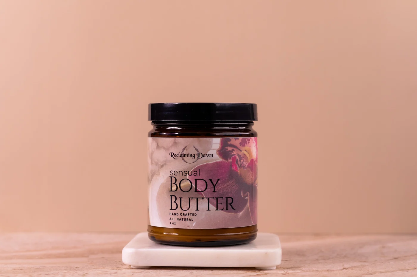 Reclaiming Dawn body butter jar on a marble riser against a warm tan backdrop