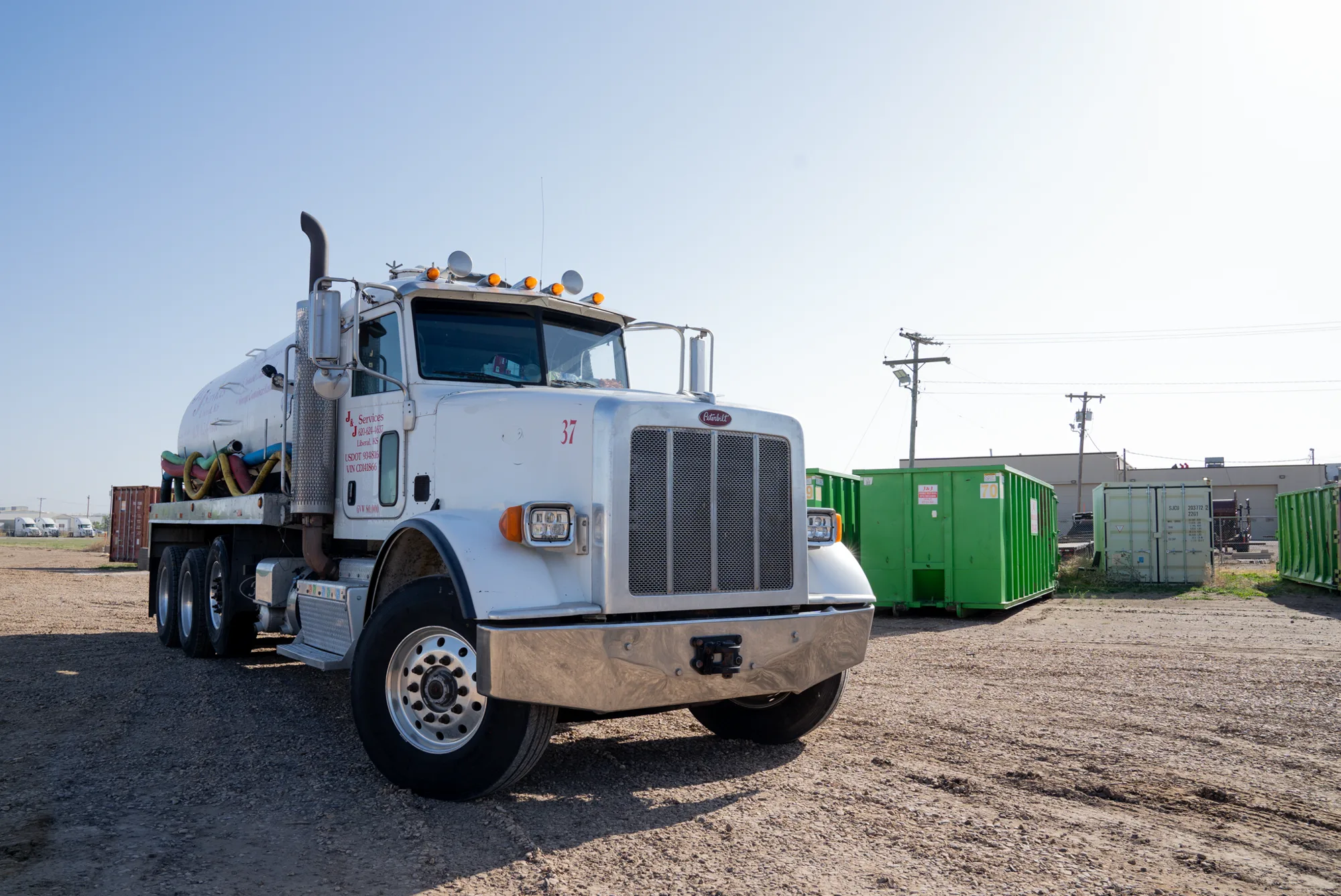 A branded J and J Services LLC vacuum truck in the yard with a row of green roll-off containers behind it